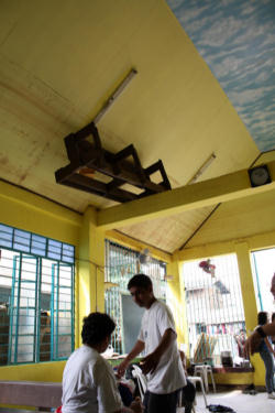 Image of 'Pew, That Was Close': Flood survivors receive aid at a chapel in Marikina, Philippines. When tropical storm Ketsana hit the island, massive flooding drove thousands from their homes. Father Javier Mexicano, shown here standing, was caught in his small parish house during the storm. He and another priest broke through the roof, waited there for the waters to settle, and eventually swam to safety.
<br>
<br>A pew is lodged in the rafters of the chapel, where it floated during the flood.The chapel's name? Our Lady of Perpetual Help.
<br>
<br>Photo: Laura Sheahen/Catholic Relief Services
<br>
<br>
