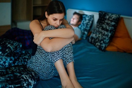 Image of Sad wife sitting on bed edge, holding her legs after fight, her husband is lying in bed in background, his face is lit by cell phone.